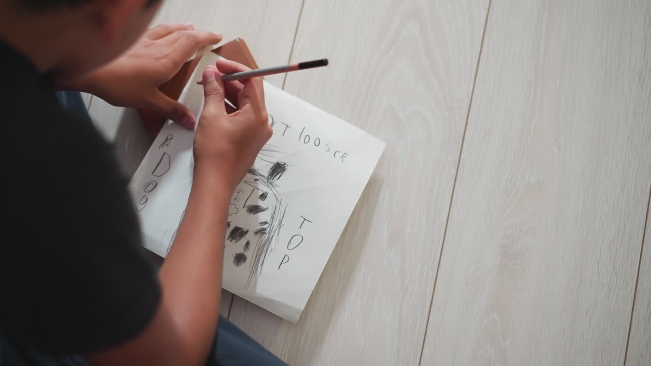 Vista posterior de un joven dibujando un gato en un papel blanco con un lápiz mientras está sentado en el suelo de madera, concentrado en dibujar un gato juguetón con detalles de texto que muestran creatividad temprana y expresión artística.
