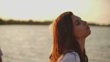 A-female-student-is-dancing-on-the-open-air-party-with-beer.-She-smiles-and-touchs-her-long-dark-hair-and-enjoys-party-time-on-the-river-coast-at-sunset.