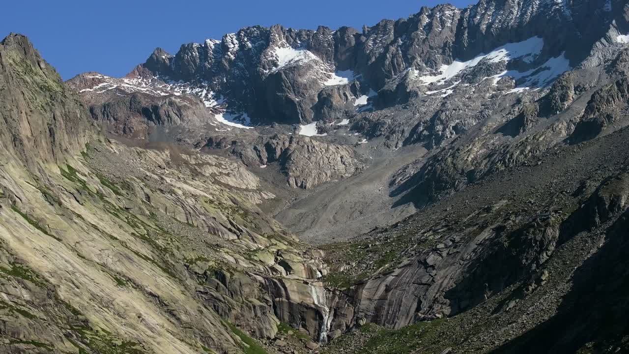 Revealing drone shot of alpine mountain range with sharp rocky snowy peaks, the steep cliffs descend into a rugged valley where a narrow waterfall flows between textured rock formations