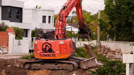 an orange excavator removes a mature palm tree from a landscaped yard in a residential gold coast neighborhood, under natural daylight with steady camera framing
