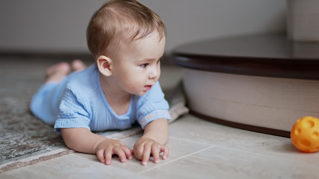 Nice cute child lies on the floor watching the ball rolling in front of him. Little boy crawling up to the toy watching it move. Close up.