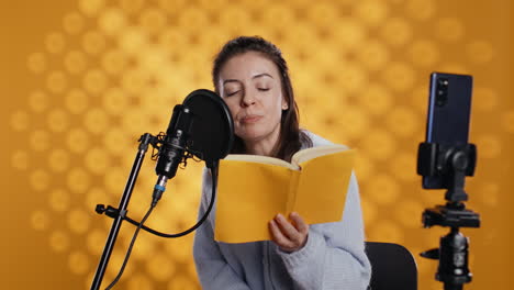 Influencer-filming-reading-book-with-smartphone-on-tripod,-studio-background