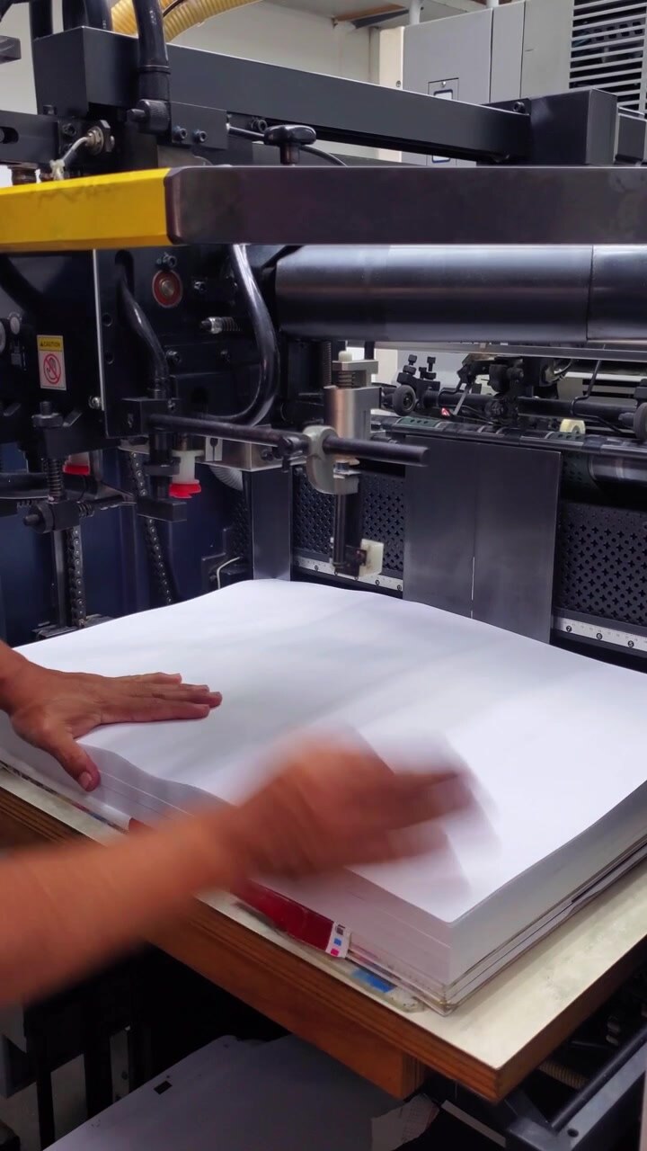 Close-up shot of a skilled worker meticulously setting up paper stacks in an offset printing machine, showcasing industrial efficiency and technical attention to detail. Vertical Video