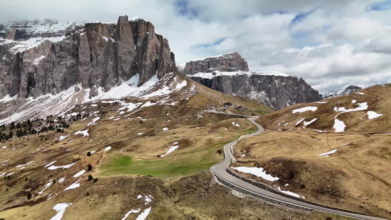 A cinematic flying backwards drone reveal of Sella Pass in the Dolomites, Italy. The scene opens with a winding alpine road and gradually unveils the dramatic, snow-dusted peaks of the Italian Alps