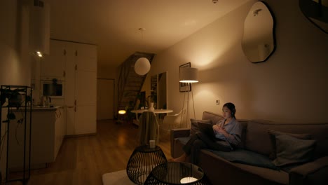 woman using a tablet on a couch in a modern living room at night.