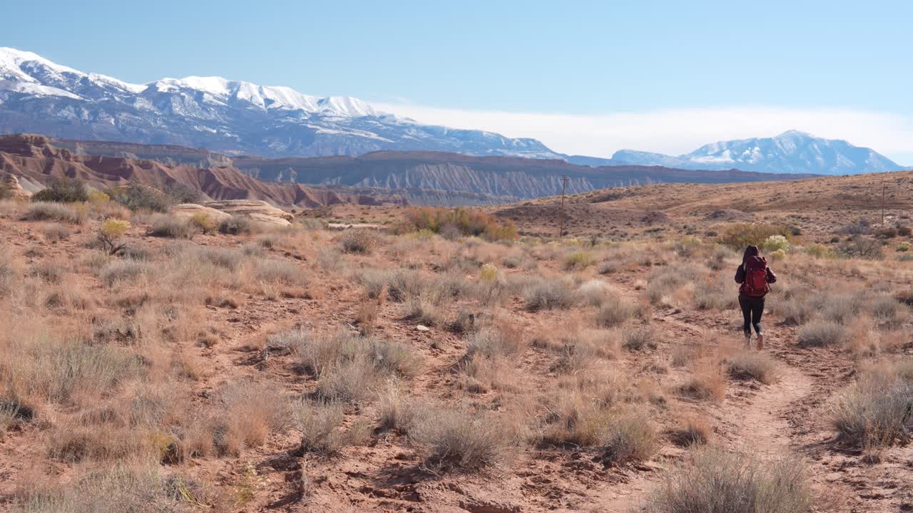 Donna solitaria con zaino che cammina su un sentiero in un paesaggio desertico con montagne innevate all'orizzonte