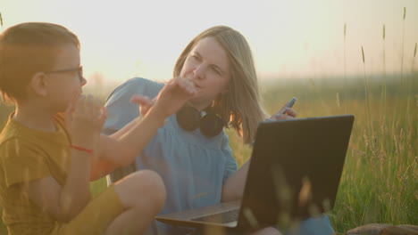 una mujer sentada en la hierba con una computadora portátil en el regazo, usando simultáneamente su teléfono. ella juega para que un niño no toque su computadora portatil