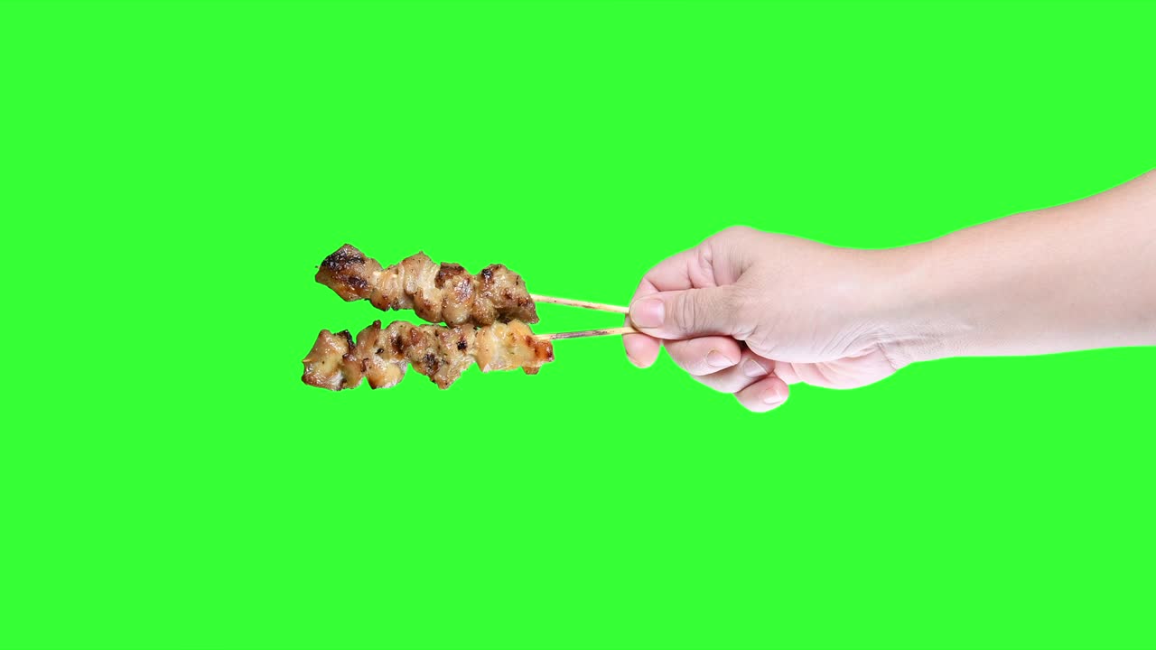 mano sosteniendo pollo satay a la parrilla en el fondo de la pantalla verde.