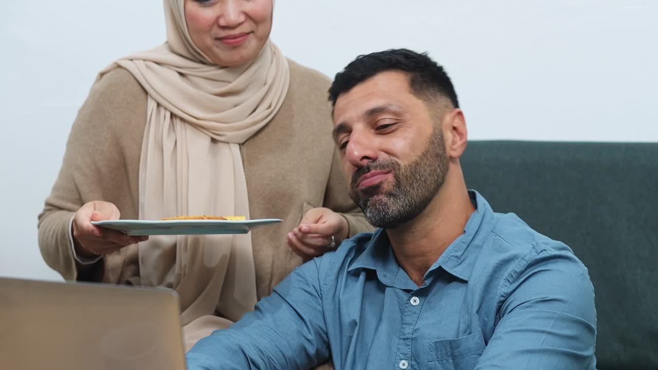 Loving Muslim wife feeds her hard working husband snacks while he works at home with laptop device