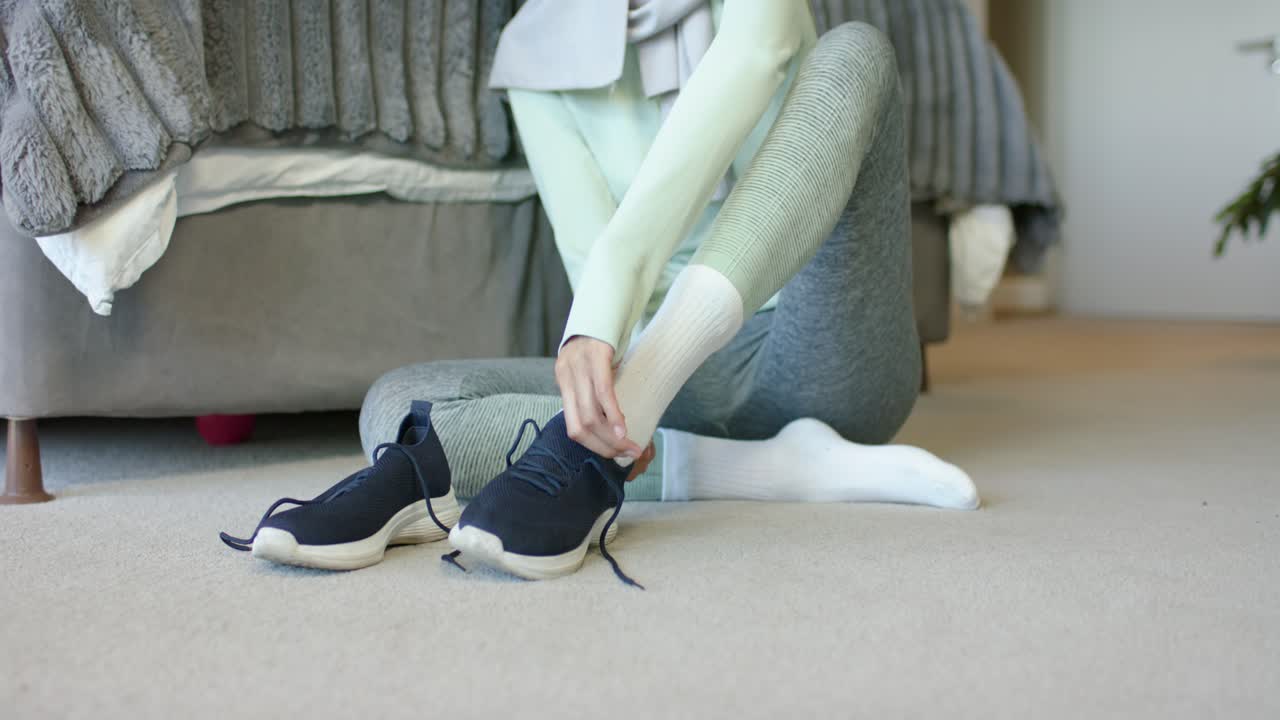 Donna mediorientale con hijab seduta in camera da letto vicino al letto, si sta mettendo delle sneakers blu scuro per uscire.