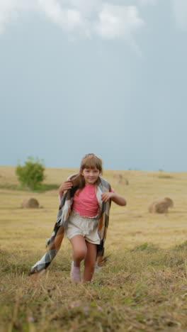 una niña feliz con una camiseta rosa y pantalones cortos corriendo a través de un vasto campo abierto con una manta sobre los hombros, usándola para cubrir la cabeza, el viento soplando, balas de heno doradas en el fondo