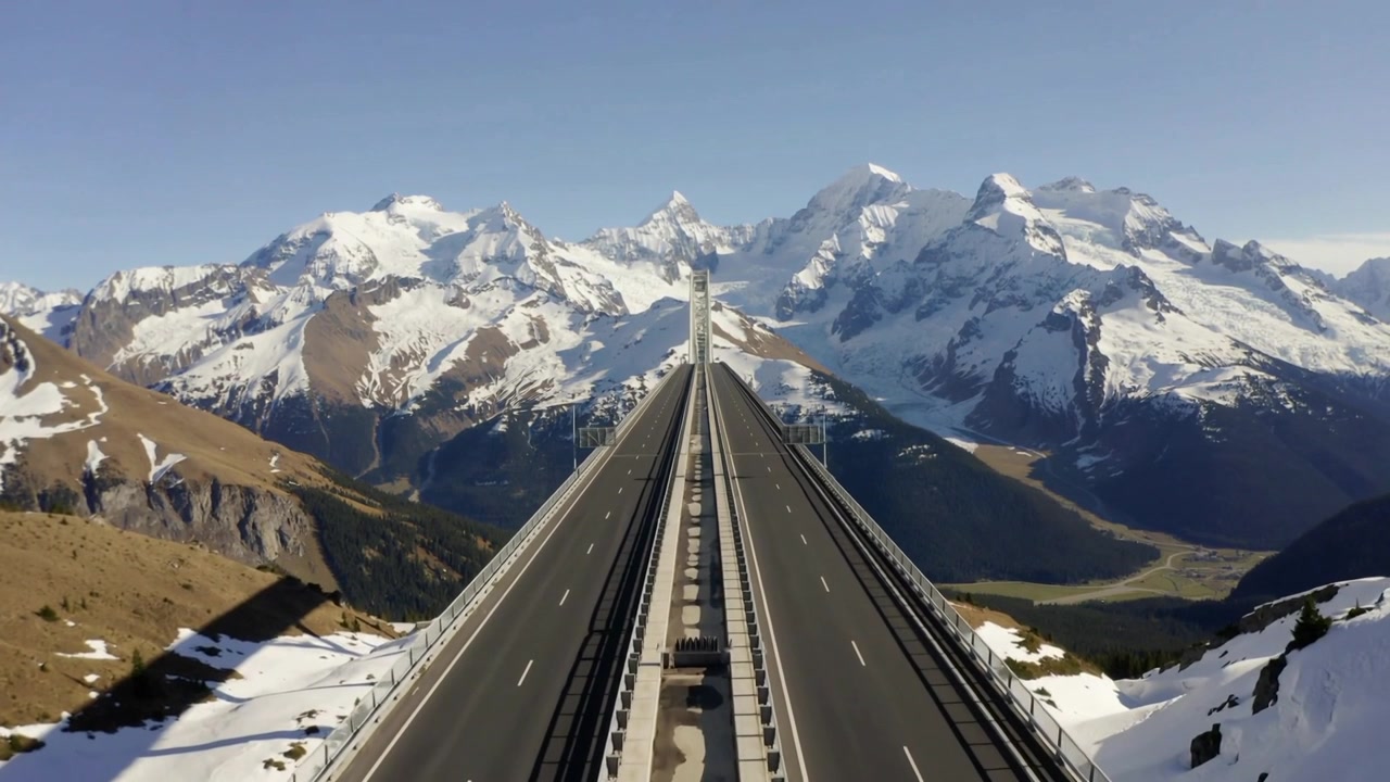 Drone in movimento che avanza lungo un alto viadotto verso cime innevate, inquadrando le vette che mostrano uno spartiacque centrale.
