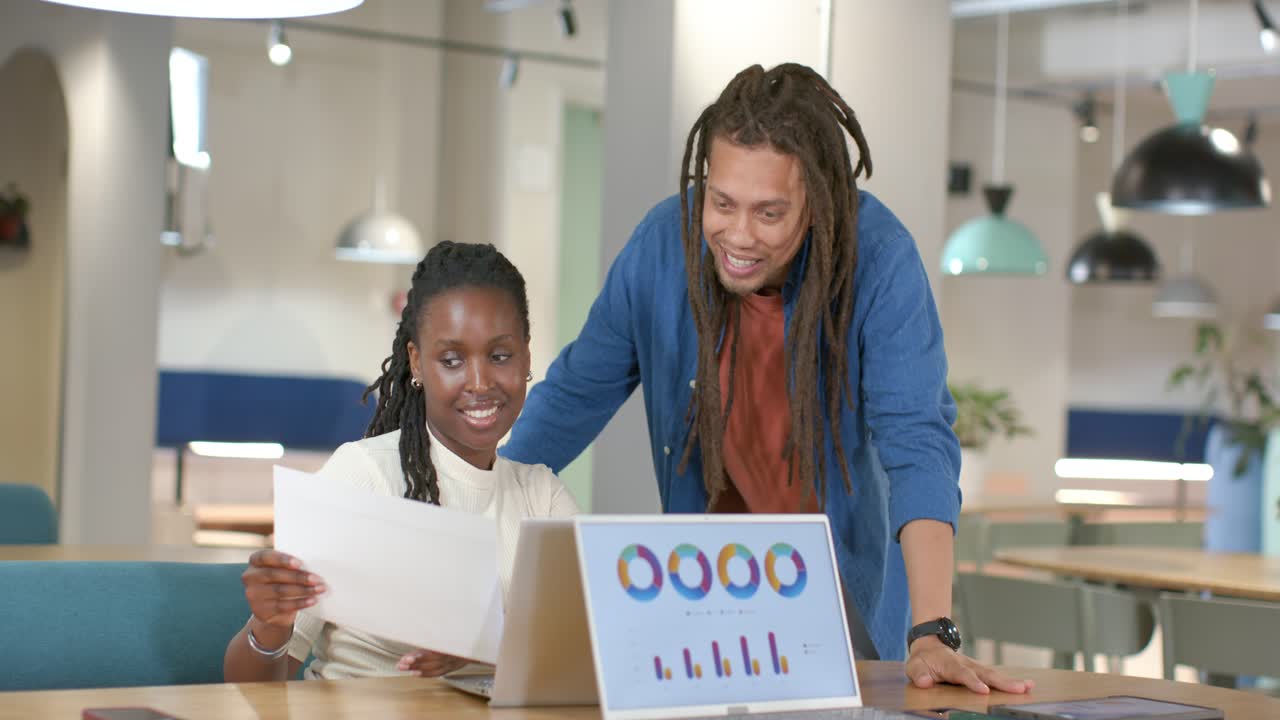 Collègues afro-américains avec un homme en chemise bleue et une femme tenant des graphiques, examinant des données sur un ordinateur portable.