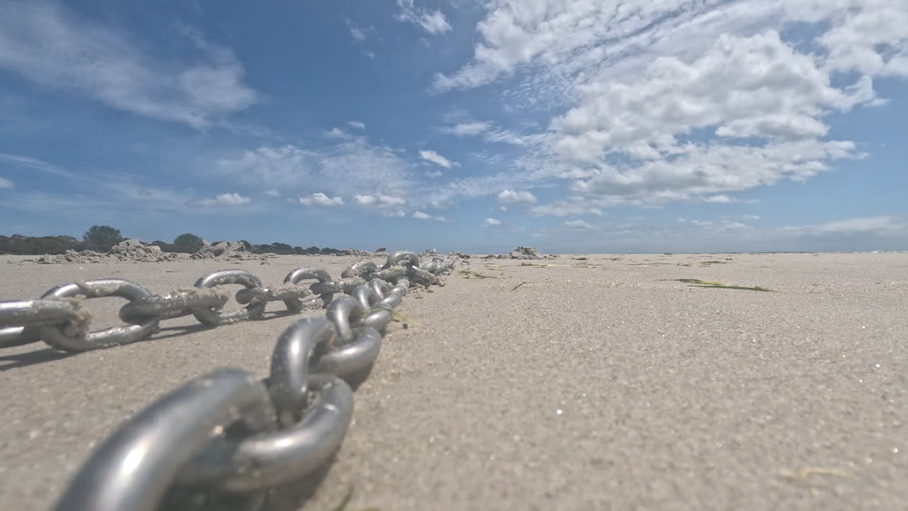 A chain beach anchor moves gently on the sandy shore under a blue sky with clouds. The scene captures the beach environment and the anchor's purpose