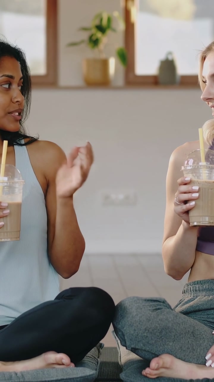 Vertical video: Sipping women glancing postworkout at home in gymwear on mats holding yellow straws