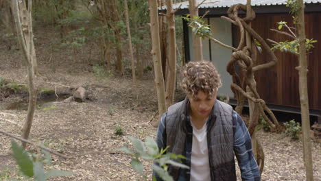 young man exploring forest near cabin, looking up at trees thoughtfully