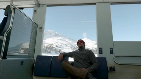 young caucasian man sittingnext to a train window and looking outside to snow covered mountains during a sunny winter day