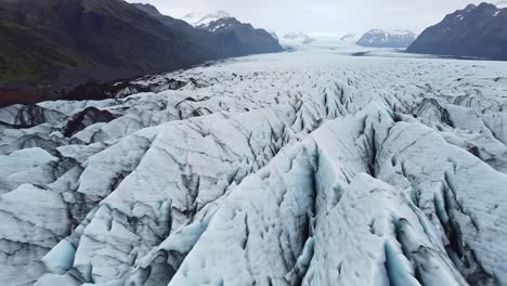 Explore the stunning expanse of a glacier in Iceland captured from above, showcasing intricate ice formations and rugged terrain under a cloudy sky during the early morning.