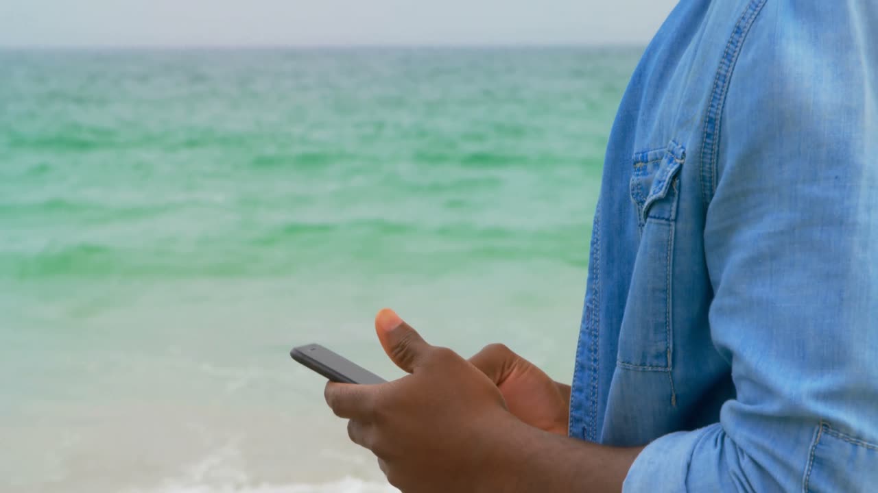 Vista laterale di uomo afroamericano che usa il telefono cellulare sulla spiaggia 4k