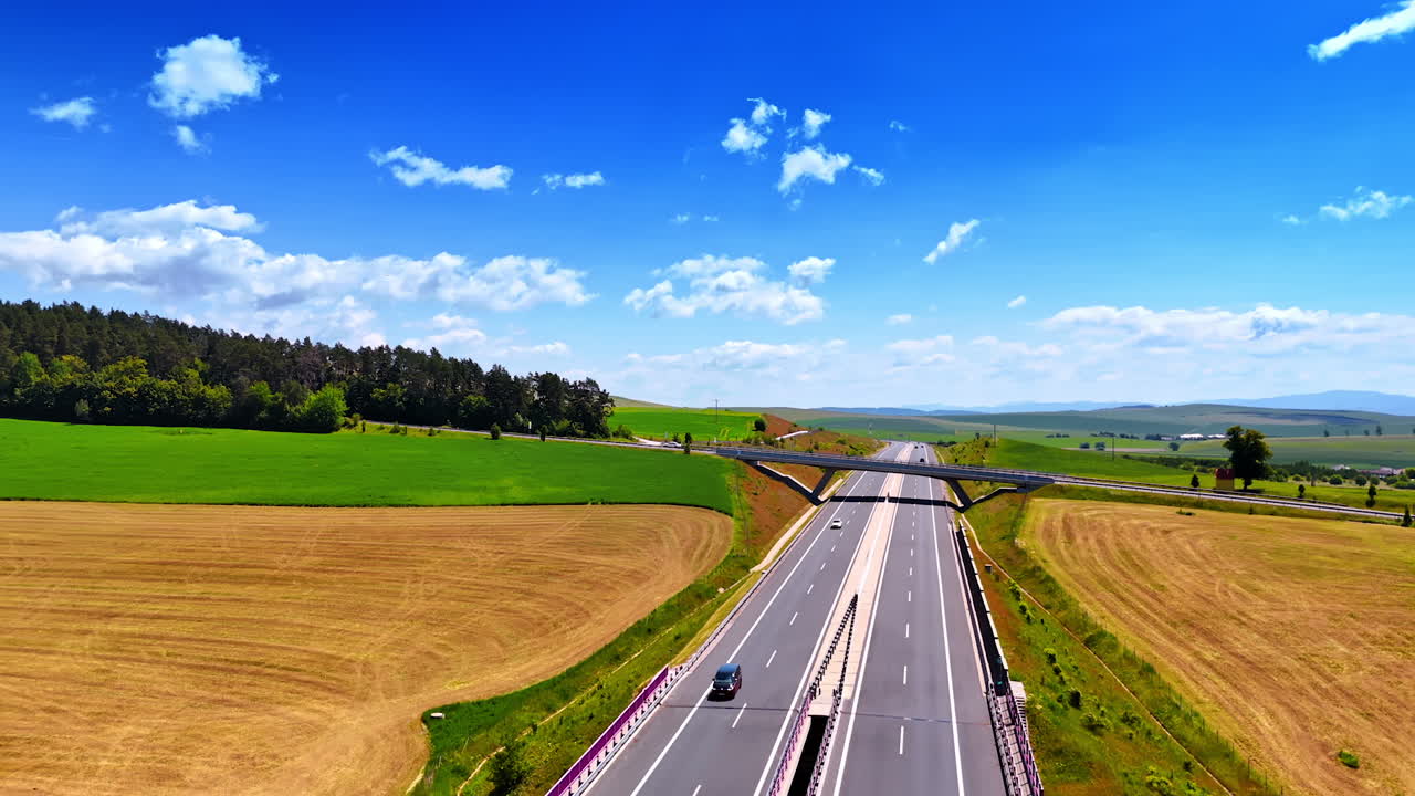 Open highway under a bright sky. Cars travel on a wide highway surrounded by green fields and a blue sky with fluffy clouds in the daytime