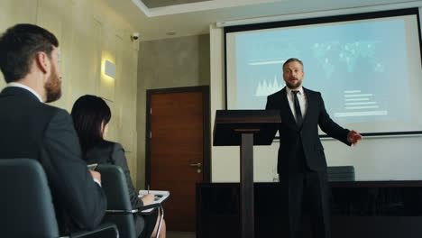 camera zoom in on caucasian businessman talking on a podium in a conference room and showing some charts and graphics on the big screen