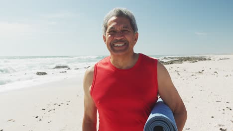 Portrait-of-hispanic-senior-man-standing-on-beach,-holding-yoga-mat-and-smiling