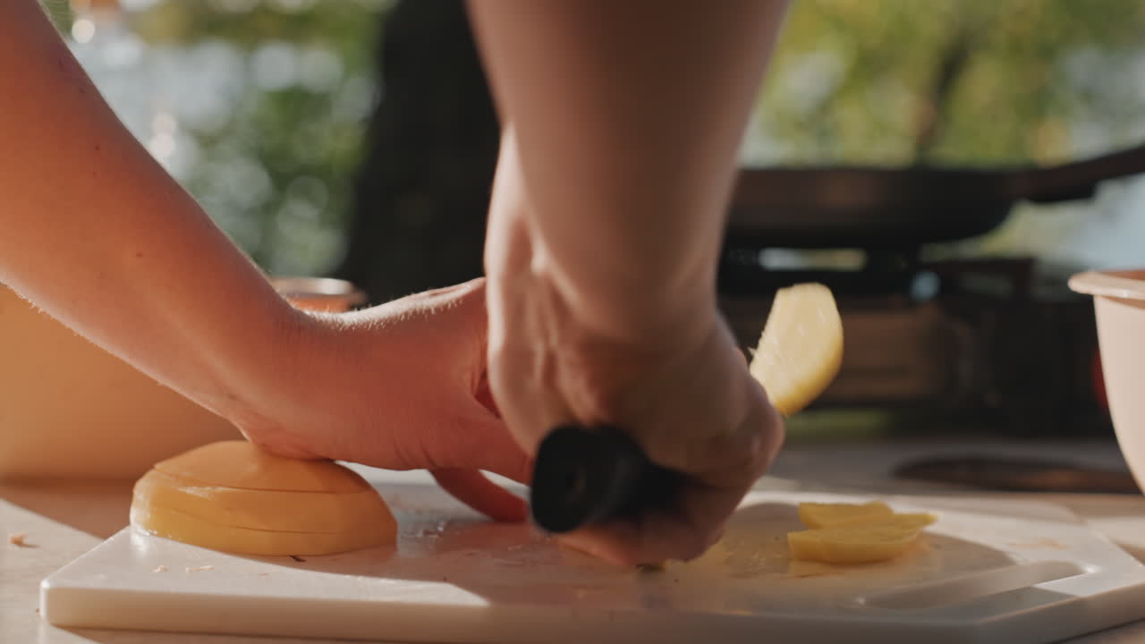 Sunlit camping cutting board hands slicing potato rounds, closeup of careful knife work on white board near portable stove, warm morning light, tranquil campground atmosphere, pot and pan blurred