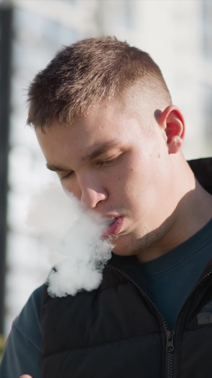 Close up of man exhaling smoke outdoors with soft lighting. Man wears black jacket and blue shirt holding object from which man smokes. background is blurred urban environment with natural