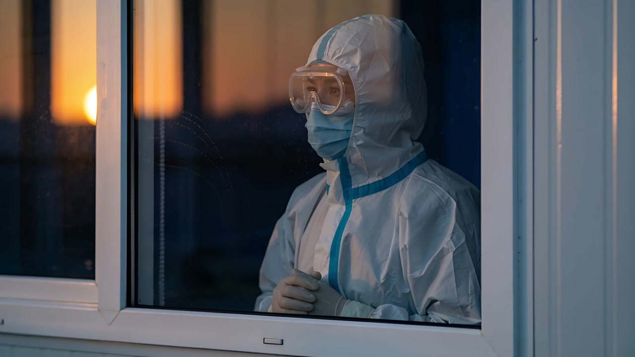 Medico in piedi con DPI incappucciato e nastro blu, che osserva il bagliore mutevole del tramonto attraverso la finestra, tenendo un oggetto.