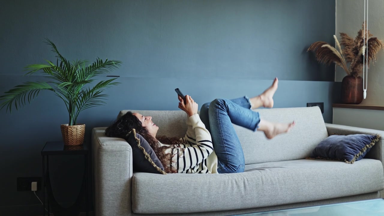 Ung kvinde slapper af på sofaen og bruger smartphone derhjemme