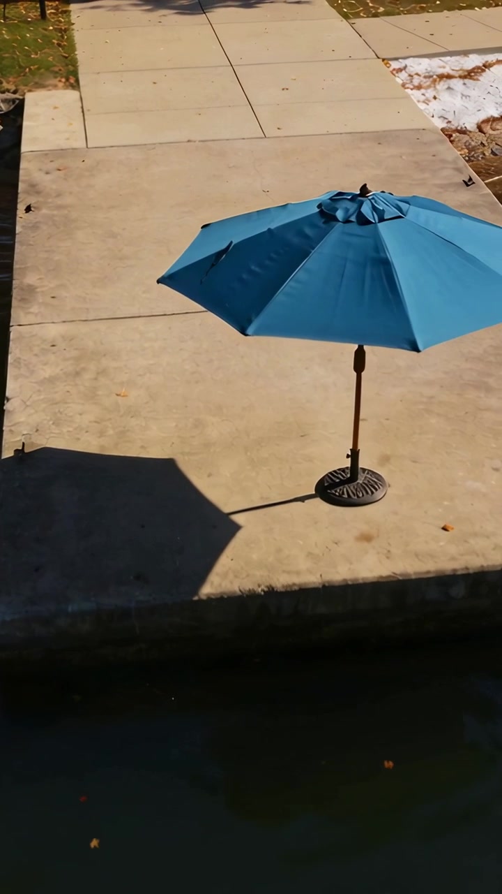 Video verticale: Panoramica della telecamera che rivela un ombrellone color verde acqua che proietta un'ombra su un patio in cemento vicino a uno stagno.