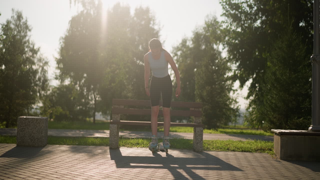 una joven con una camiseta azul claro intentando equilibrarse en patines mientras se levanta de un banco en un parque soleado, con zapatillas blancas debajo del banco y el fondo iluminado por la luz del sol