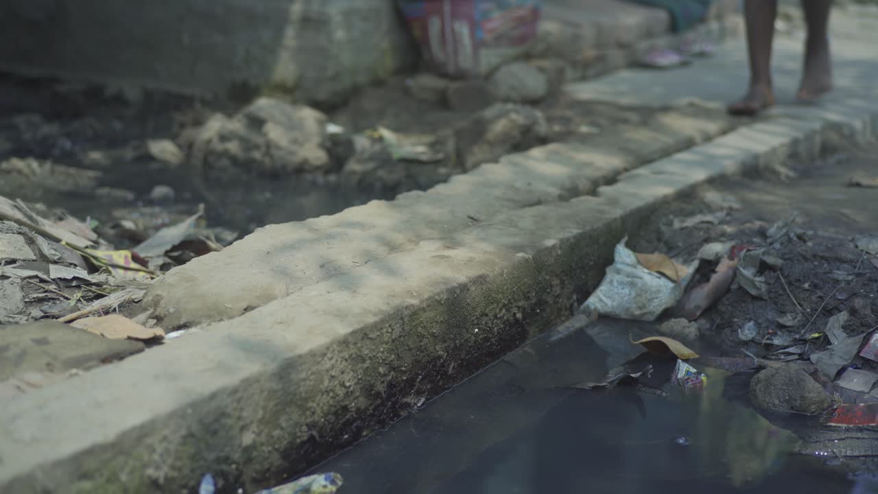 젊은 인도의 가난한 고아 소녀, 시골 마을의 쓰레기 근처 배수구에 벌거벗은 더러운 발을 들고 차선을 걷고 있습니다.