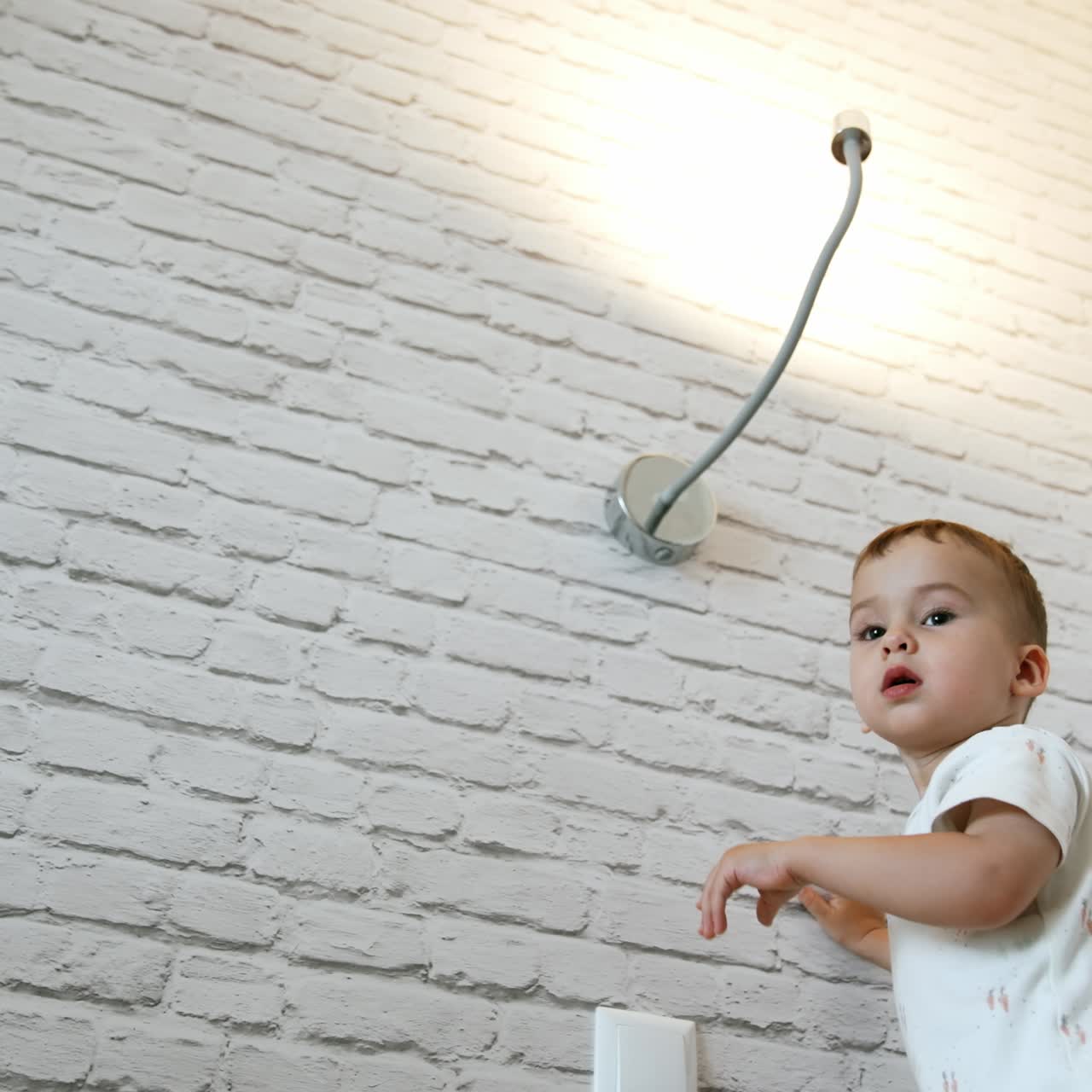 Little baby boy playing with light at home. Child stands at the wall reaching to the lamp. Low angle view