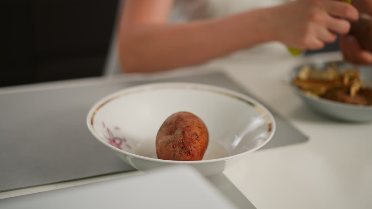 Close up of woman lifting potato from bowl while shaking off excess water in preparation for peeling, with one potato already in dish on table during meal preparation in clean kitchen setting