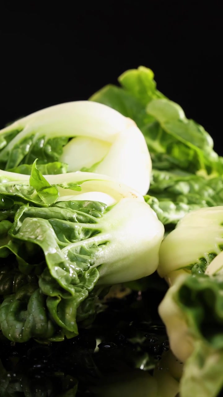 Whole bok choy vegetables fall and settle on a reflective, wet black surface under dramatic studio lighting, highlighting freshness and texture with minimal camera movement