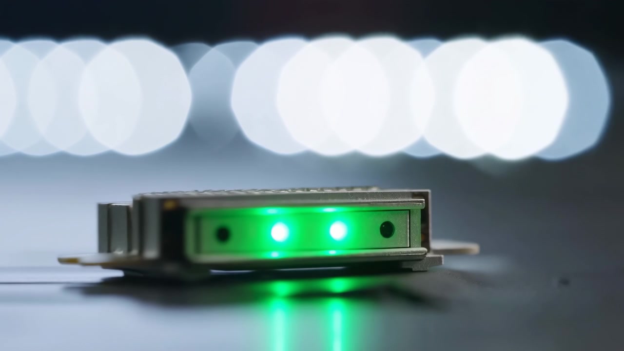 Ein grün beleuchtetes rechteckiges Objekt liegt auf einer reflektierenden Oberfläche, während helle Punkte entlang seiner Vorderkante leuchten, mit sanften Hintergrundlichtern, die eine geringe Tiefenschärfe erzeugen, und kühler Innenbeleuchtung um den Rahmen herum.