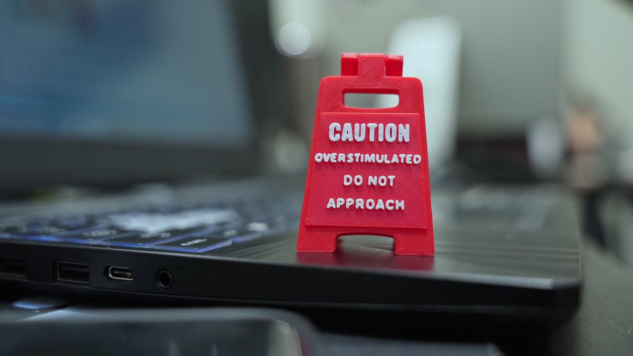 Close-up of a red "Caution Overstimulated" sign on a laptop keyboard in a workspace. A blurred hand works in the background, illustrating office burnout and mental health awareness.