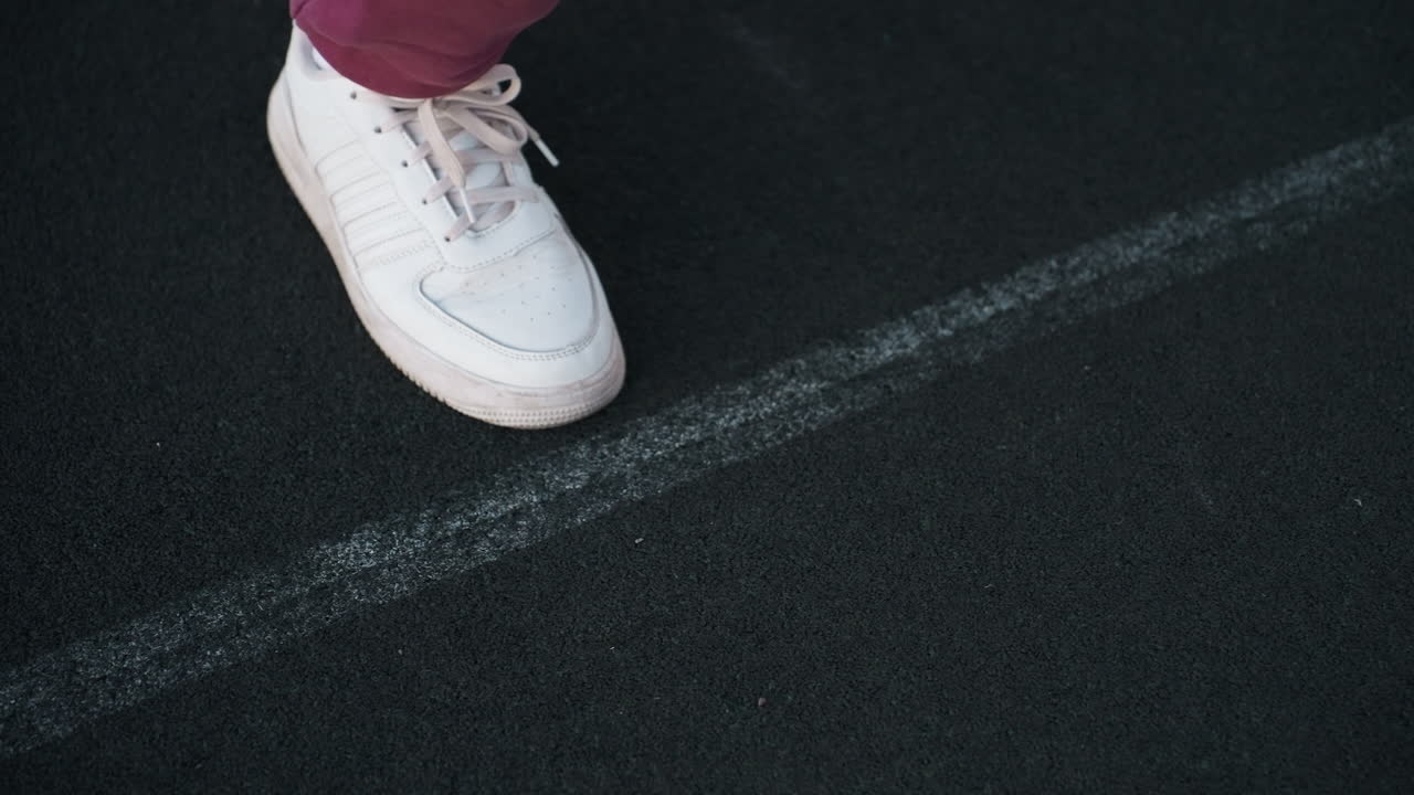 Leg view of fitness instructor placing foot on court line with hands positioned on black asphalt court near white barrier topped with chain link fence wearing maroon tracksuit and white sneakers