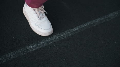 leg view of fitness instructor placing foot on court line with hands positioned on black asphalt court near white barrier topped with chain link fence wearing maroon tracksuit and white sneakers