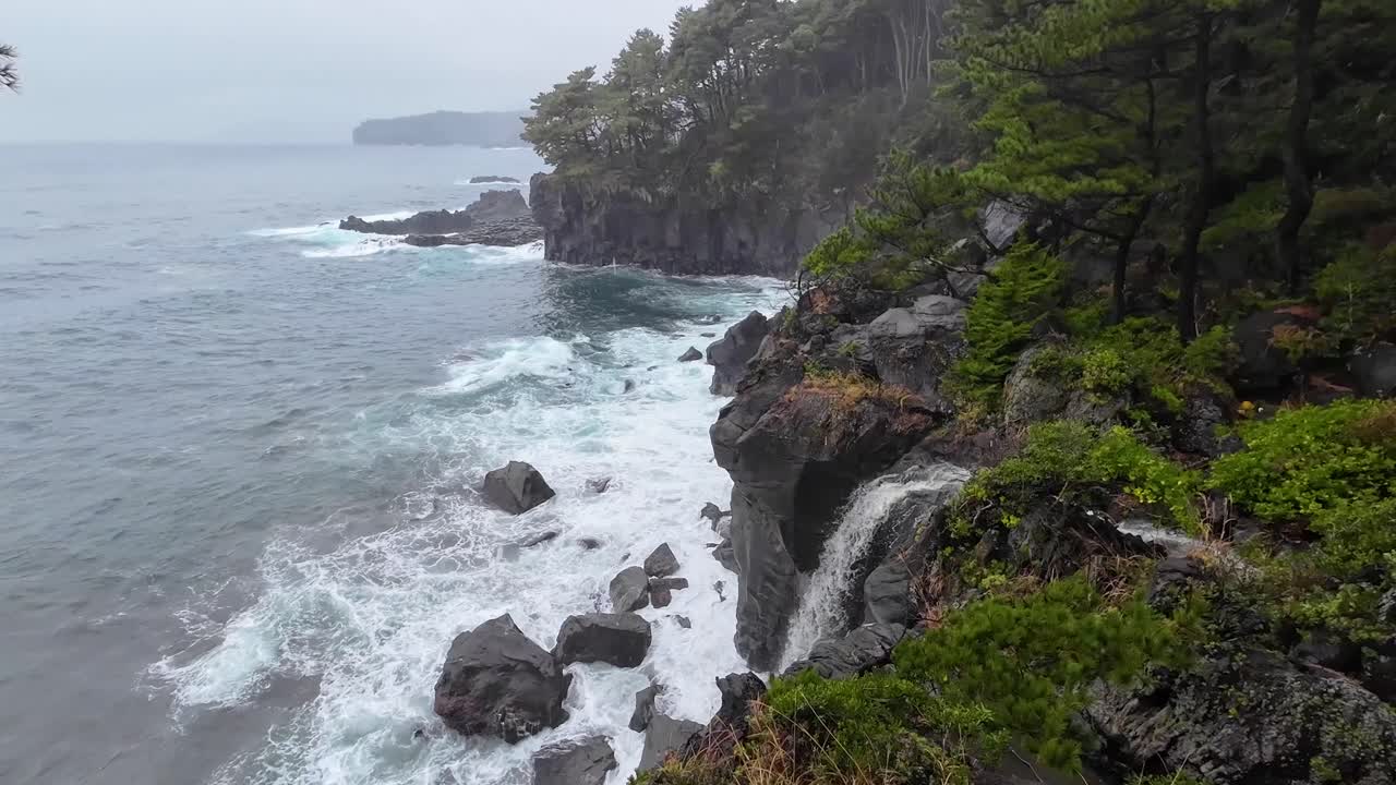 Dramatic coast line with strong waves crashing along the cliffs overlooking the sea and ocean from the forest side and a waterfall coming from the river falling into the ocean