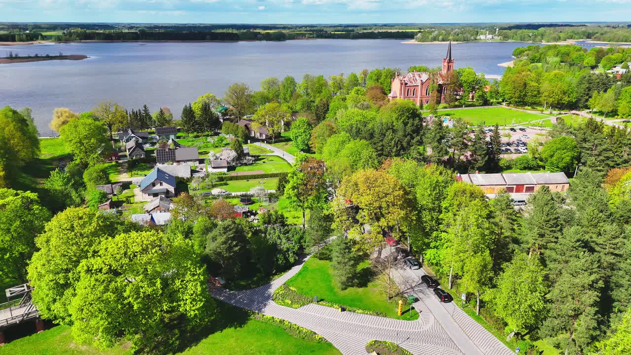 A peaceful riverside town with a prominent red-brick church, tree-lined roads, and scattered houses viewed from above. Lush spring foliage enhances the scenic landscape.