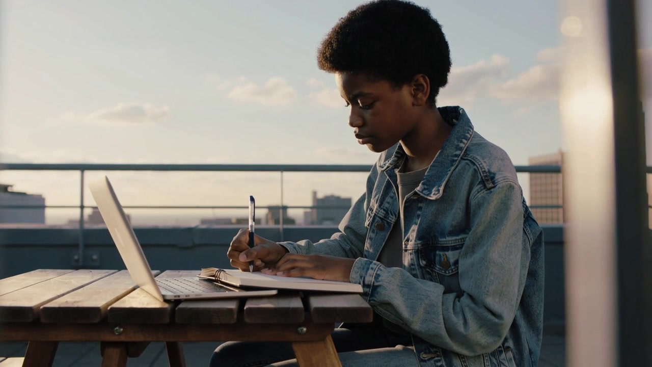 Estudante sentado chegando, de jaqueta jeans escrevendo anotações em uma mesa no terraço, estudando com o laptop à esquerda