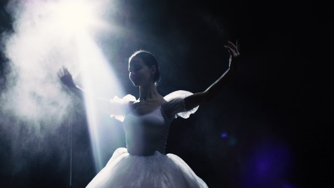 Mid Shot of a Beautiful Young Ballerina Spinning Gracefully in the Spotlight. Powder and Smoke Shine in the Darkness Around Her. She's Wearing White Tutu Dress that Sparkles in the Light. In Slow Motion.