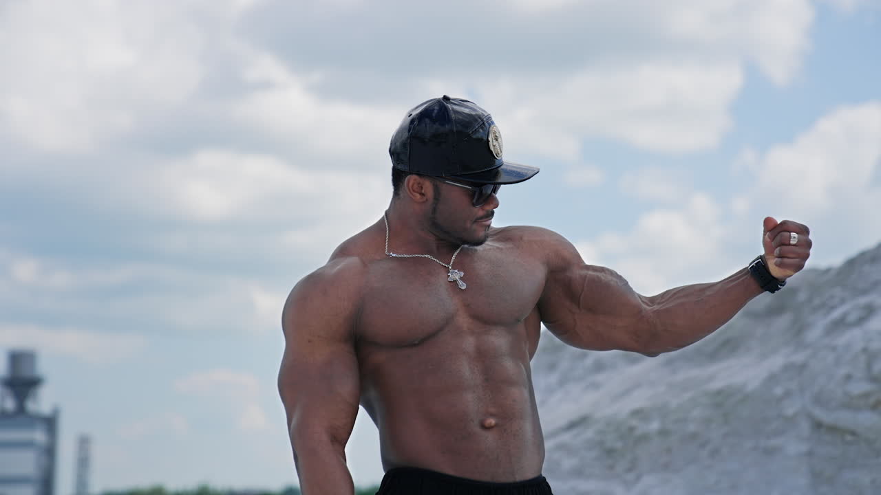 Portrait of muscular athlete in the mountains. Shirtless african american sportsman in black cap and sunglasses showing his arms muscles outdoors.