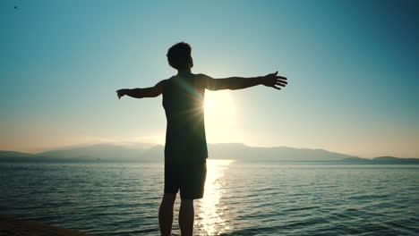 lonely man standing in front of lake with his arms open enjoying sunset