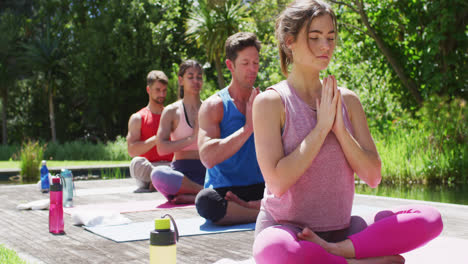 diverse male and female group practicing yoga sitting in a row with eyes closed in sunny park