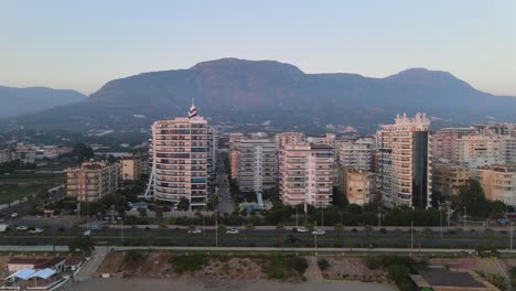 Toma-Aérea-De-Una-Fila-De-Edificios-Residenciales-Y-Hoteles-Junto-Al-Mar-En-Alanya,-Turquía,-Con-La-Vista-De-La-Cordillera-En-El-Fondo