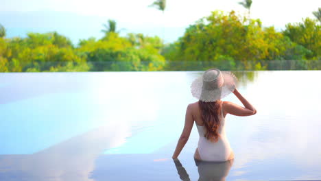 A-woman-with-her-back-to-the-camera-sits-along-the-edge-of-a-swimming-pool-reaches-up-to-adjust-her-sunglasses