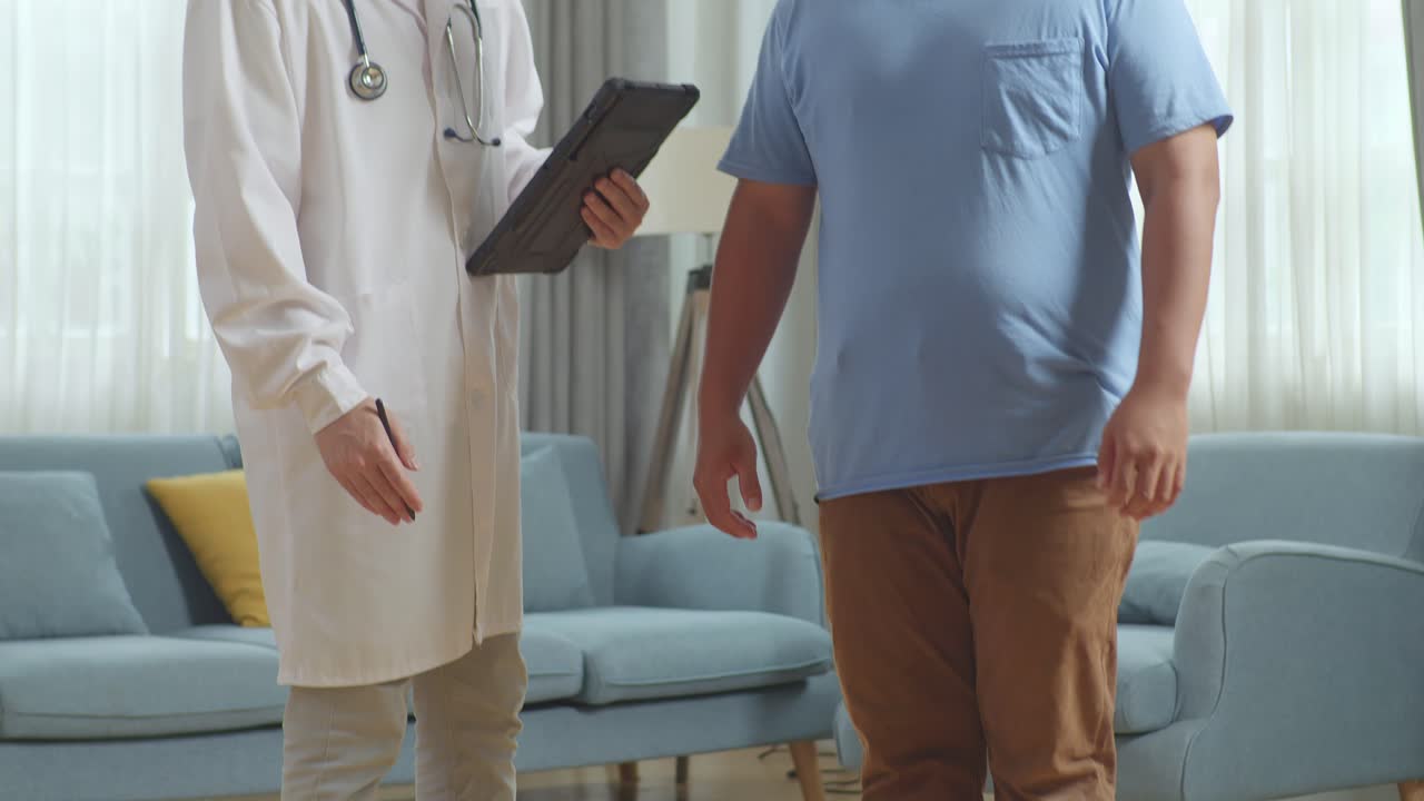 Asian Male Doctor With A Fat Male Patient In Clinic. The Patient Measuring Weight Using Weighing Machine, The Doctor Recording On A Tablet